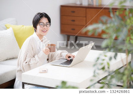 A woman wearing glasses drinking coffee and using a computer in a living room with houseplants, looking at the camera 132426861