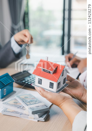 Close up of real estate agent handing house key to client with contract paperwork, calculator and house models on desk, concept of home ownership, mortgage loan, property investment and success. 132428279