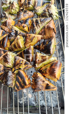 Grilled banana leaf parcels on charcoal grill, smoky savory aroma and golden charred texture evokes street food comfort 132428559