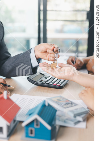 Close up of real estate agent handing house key to client with contract paperwork, calculator and house models on desk, concept of home ownership, mortgage loan, property investment and success. Close up of real estate agent handing house key to client with contract paperwork, calculator and house models on desk, concept of home ownership, mortgage loan, property investment and success. 132428939