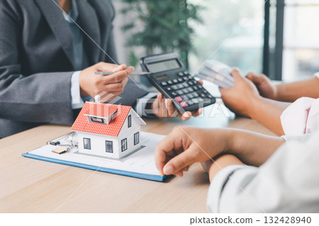 Close up of real estate agent handing house model to client with calculator and contract paperwork on desk, concept of mortgage loan, home ownership, property investment and housing agreement. Close up of real estate agent handing house model to client with calculator and contract paperwork on desk, concept of mortgage loan, home ownership, property investment and housing agreement. 132428940