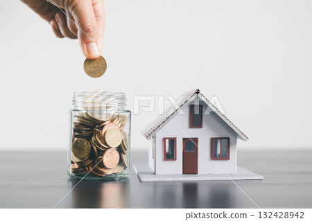 Hand putting coin into glass jar full of money beside house model, symbolizing savings, mortgage loan, real estate investment, property finance, home ownership and wealth management concept. Hand putting coin into glass jar full of money beside house model, symbolizing savings, mortgage loan, real estate investment, property finance, home ownership and wealth management concept. 132428942