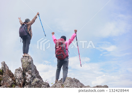 Two hikers with backpacks raising arms and trekking poles on rocky mountain peak, symbol of adventure, achievement, teamwork, freedom, success, exploration and outdoor lifestyle inspiration. Two hikers with backpacks raising arms and trekking poles on rocky mountain peak, symbol of adventure, achievement, teamwork, freedom, success, exploration and outdoor lifestyle inspiration. 132428944
