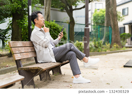 一名中年男子坐在長椅上操作智慧型手機 一名中年男子坐在長椅上操作智慧型手機 132430329