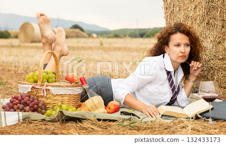 Woman in white blouse in office clothes, lying on blanket and reading book. Picnic on farm field 132433173