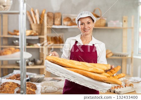 Happy woman baker with joyful expression holds a tray of freshly baked baguettes in bakery shop, showcasing the concept of the food industry and catering services Happy woman baker with joyful expression holds a tray of freshly baked baguettes in bakery shop, showcasing the concept of the food industry and catering services 132433400