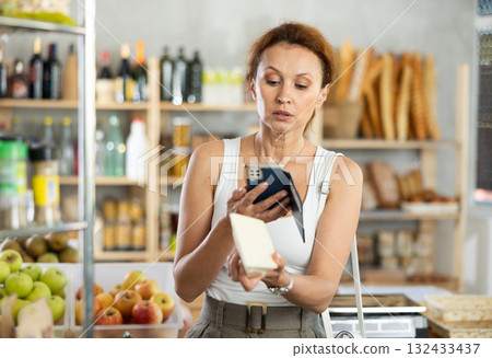 Female shopper scans QR code on cheese label using smartphone. Checking products in supermarket 132433437