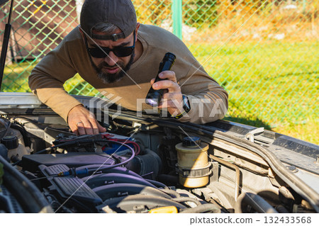 Man inspecting car engine with flashlight in backyard during daylight 132433568
