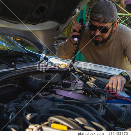 Man checking car engine with flashlight in outdoor setting Man checking car engine with flashlight in outdoor setting 132433570