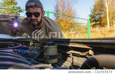 Man inspecting car engine with flashlight in autumn setting 132433573