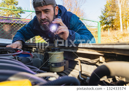 Man inspecting car engine with flashlight in sunny outdoor setting Man inspecting car engine with flashlight in sunny outdoor setting 132433574