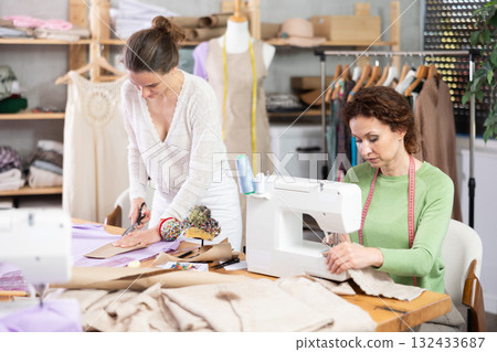Female tailor sews on a sewing machine while another tailor cuts out patterns for dress with scissors Female tailor sews on a sewing machine while another tailor cuts out patterns for dress with scissors 132433687