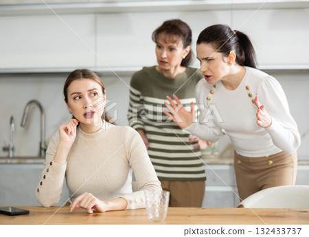 Upset middle-aged woman sitting at the kitchen table with her back to women quarreling to her Upset middle-aged woman sitting at the kitchen table with her back to women quarreling to her 132433737