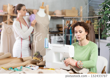 Woman tailor at work in cloth design shop. Companion work in background with mannequin 132433787