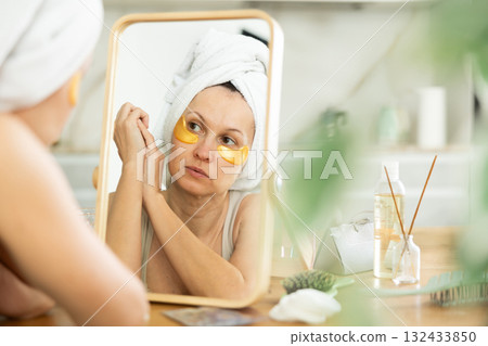 Woman in towel turban applying golden collagen eye patches Woman in towel turban applying golden collagen eye patches 132433850