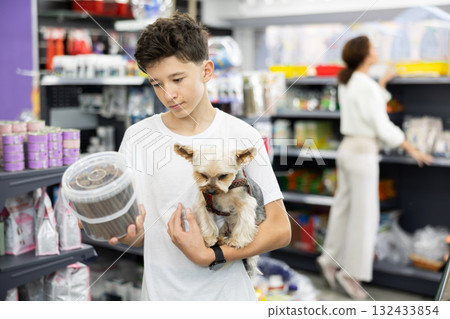 Boy with dog Yorkshire terrier in arms and read composition of product at packaging of dog dry food Boy with dog Yorkshire terrier in arms and read composition of product at packaging of dog dry food 132433854