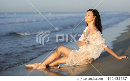 Portrait of woman in white mesh cape with glass of champagne posing on seashore Portrait of woman in white mesh cape with glass of champagne posing on seashore 132433887