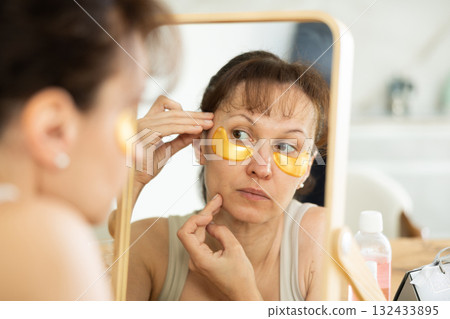 Middle-aged woman applying eye patch mask on her face sitting in front of the mirror Middle-aged woman applying eye patch mask on her face sitting in front of the mirror 132433895