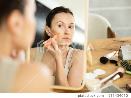 Woman applying orange concealer on cheekbone in front of mirror Woman applying orange concealer on cheekbone in front of mirror 132434050