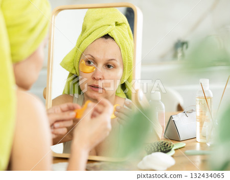 Middle-aged woman applying eye patch mask on her face sitting in front of the mirror Middle-aged woman applying eye patch mask on her face sitting in front of the mirror 132434065