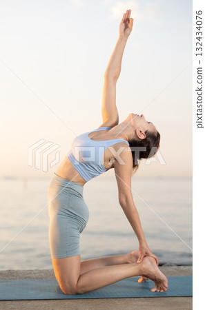 Near sea during sunset, girl performs camel pose 132434076