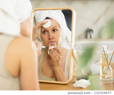 Middle-aged woman cleaning her facial skin with cotton disks sitting in front of the mirror Middle-aged woman cleaning her facial skin with cotton disks sitting in front of the mirror 132434077