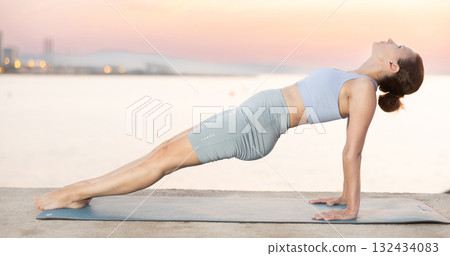 Sporty energetic experienced young woman yoga trainer performing upward plank pose on rug on beach at sunrise. Fitness concept 132434083