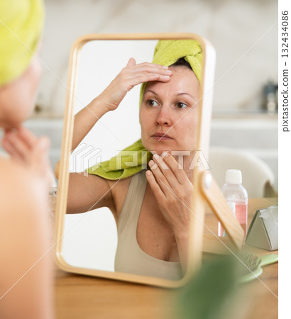 Woman in towel turban on head examining facial skin in mirror Woman in towel turban on head examining facial skin in mirror 132434086