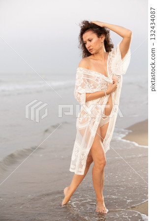 Woman in white negligee stands on coast 132434087