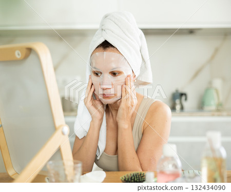 Middle-aged woman applying tissue mask on her facial skin sitting in front of the mirror 132434090