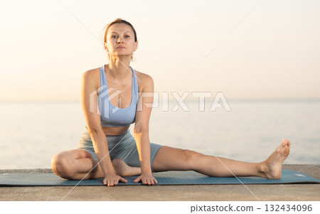 Near sea during sunset, girl performs variations of Janu Shirshasana 132434096