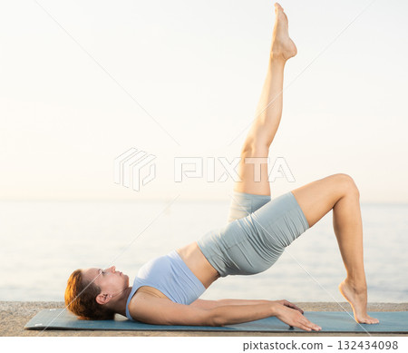Sporty female wearing sportswear standing legs up in Salamba Sarvangasana exercise during yoga on seashore at sunrise 132434098