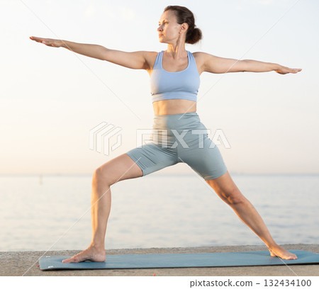 Happy woman coach standing yoga pose and making full body stretching workout on seashore at sunrise 132434100