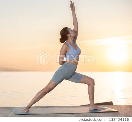 Active experienced woman teacher performs yoga asana on mat on seashore at sunset. Concept of healthy lifestyle Active experienced woman teacher performs yoga asana on mat on seashore at sunset. Concept of healthy lifestyle 132434116