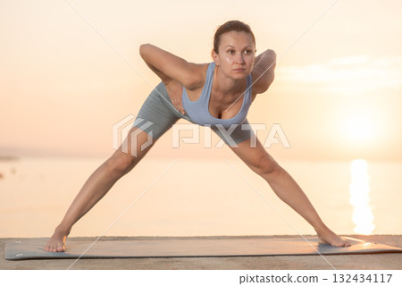 Diligent woman standing and practicing forward bend pose of yoga on mat on seashore at sunrise 132434117