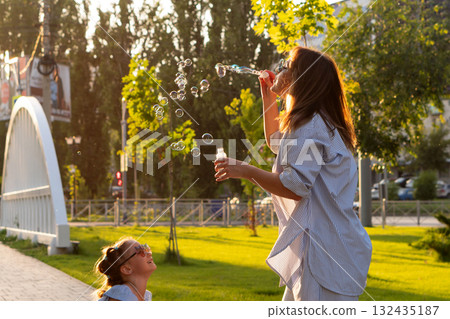 Mother blows soap bubbles as daughter watches laughing Mother blows soap bubbles as daughter watches laughing 132435187
