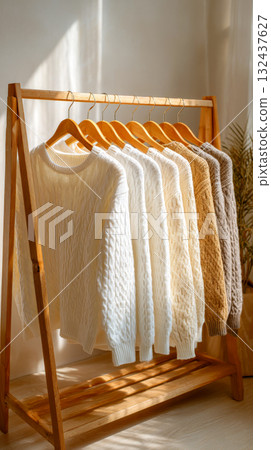Seven cable-knit sweaters in varying shades of white, cream, and brown hang neatly on wooden hangers on a minimalist wooden clothing rack 132437627