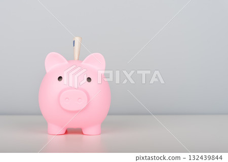 A pink piggy bank with a banknote inserted stands on the table. A pink piggy bank with a banknote inserted stands on the table. 132439844