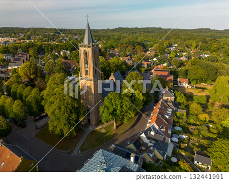 Aerial view of town with central Gothic style church, pointed steeple, red and gray rooftops, tree lined streets, and parked cars blending history with modern life. Aerial view of town with central Gothic style church, pointed steeple, red and gray rooftops, tree lined streets, and parked cars blending history with modern life. 132441991