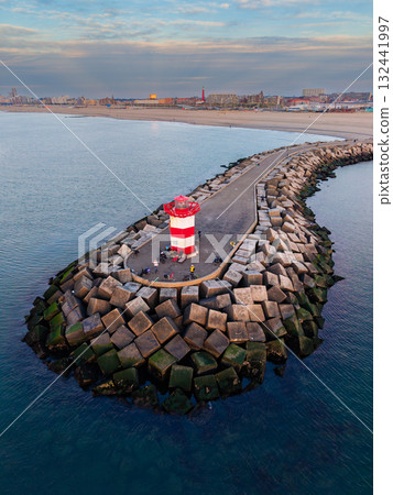 Aerial view of breakwater with red white lighthouse, people gathered near edge, concrete blocks protect harbor, coastal city and second tower in background under partly cloudy evening sky. 132441997