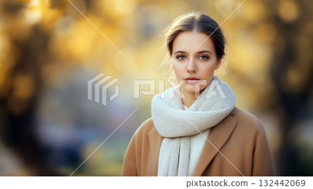 Portrait of a young woman in a beige coat and scarf standing outdoors in soft autumn light with golden bokeh background 132442069