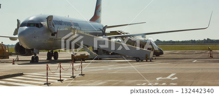 This image depicts a commercial airplane parked on the apron at an airport. The aircraft's boarding stairs are extended, waiting for passengers to embark or disembark. To the left of the stairs, we 132442340