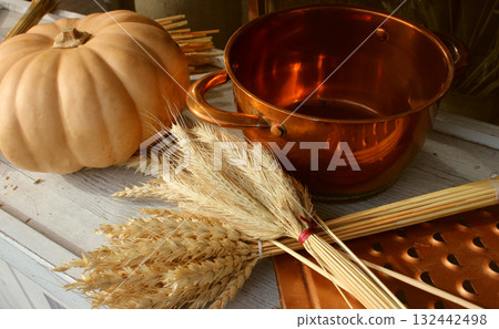 Stock photo of an autumn harvest with a bright ripe pumpkin, sheaves of harvested wheat, and brass kitchen utensils 132442498