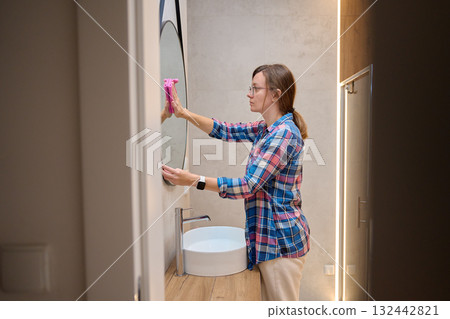Woman wearing glasses cleaning round mirror with pink cloth near sink in modern bathroom. Concept of home cleaning, hygiene, daily routine and household chores 132442821