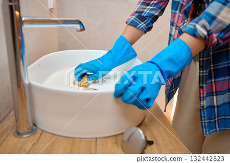 Woman wearing blue gloves cleaning cleaning white ceramic sink with sponge and soap foam in bathroom. Concept of hygiene, sanitation, household chores and daily home maintenance Woman wearing blue gloves cleaning cleaning white ceramic sink with sponge and soap foam in bathroom. Concept of hygiene, sanitation, household chores and daily home maintenance 132442823