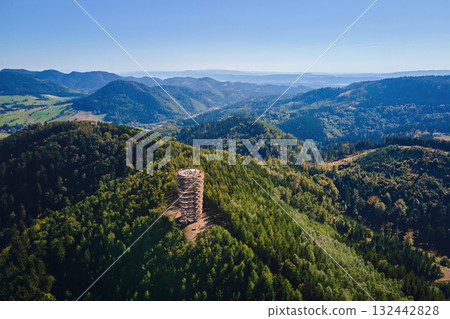 Aerial panoramic view of observation tower on forested mountain hill. Natural vast landscape with forest and distant ridges Aerial panoramic view of observation tower on forested mountain hill. Natural vast landscape with forest and distant ridges 132442828