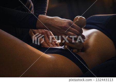 A professional therapist performing a wood therapy (maderotherapy) massage using a wooden roller on a woman's legs, belly and glutes A professional therapist performing a wood therapy (maderotherapy) massage using a wooden roller on a woman's legs, belly and glutes 132443113