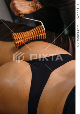 A professional therapist performing a wood therapy (maderotherapy) massage using a wooden roller on a woman's legs, belly and glutes 132443124