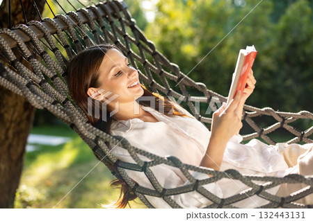 Young woman lying in hammock, reading a book and looking contented Young woman lying in hammock, reading a book and looking contented 132443131