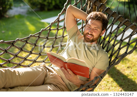 Caucasian bearded young man lying in hammock with a book in hands Caucasian bearded young man lying in hammock with a book in hands 132443147